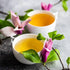 Two cups of Magic Hour Magnolia Oolong Tea with a light golden hue sit on a textured gray surface, surrounded by green leaves and vibrant purple-pink flowers, creating a serene and inviting setting.
