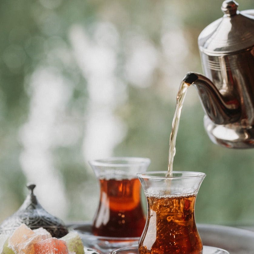 A close-up of two traditional glass cups filled with Magic Hour&#39;s Tea of The Rising Sun : Japanese Breakfast on a tray. A silver teapot is pouring tea into one of the cups. In the foreground, there are pieces of colorful Turkish delight. The background is blurred, showing green foliage.