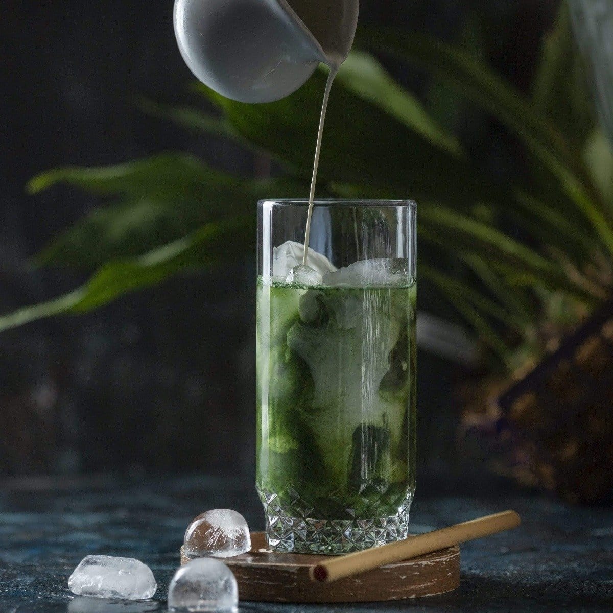 A tall glass filled with a green beverage and ice sits on a wooden coaster. A small pitcher pours a creamy liquid into the glass. A bamboo straw rests beside the coaster. The background features green foliage and a dark, moody ambiance, perfect for savoring Organic Ceremonial Matcha 700 from Club Magic Hour.
