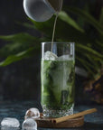A tall glass filled with a green beverage and ice sits on a wooden coaster. A small pitcher pours a creamy liquid into the glass. A bamboo straw rests beside the coaster. The background features green foliage and a dark, moody ambiance, perfect for savoring Organic Ceremonial Matcha 700 from Club Magic Hour.