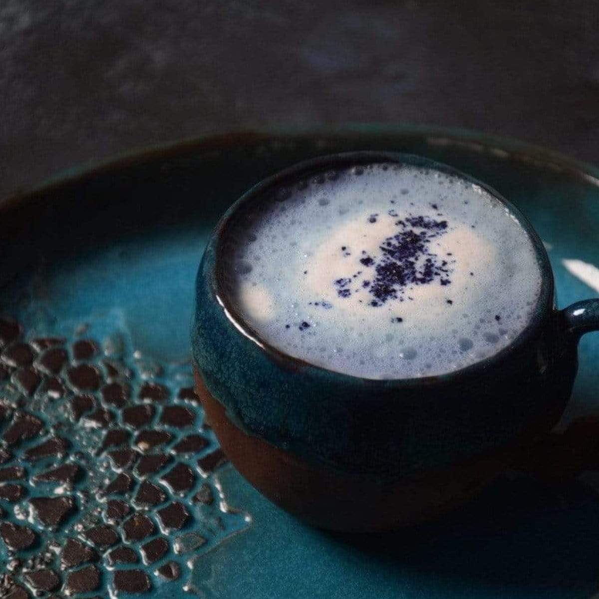 A cup in deep blue ceramic from the "Butterfly Pea Flower: Finely Cut- ARCHIVE" collection sits on a matching tray, brimming with a frothy drink lightly dusted with dark powder. The tray's intricate pattern enhances its elegant and artistic appeal.