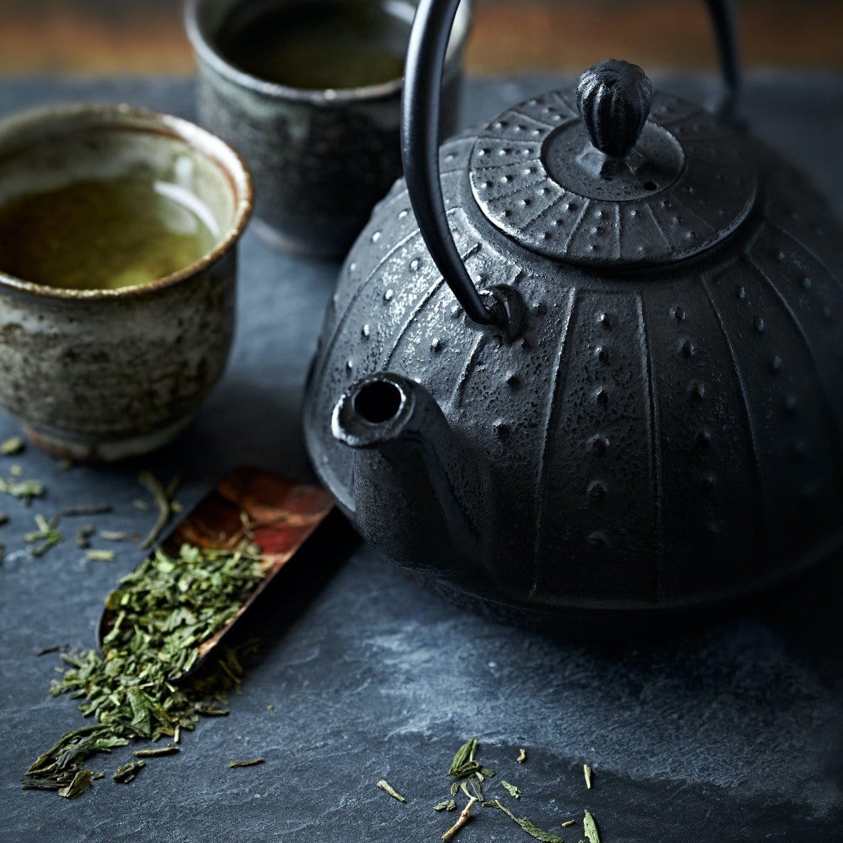 A close-up of a black, ornate cast iron teapot with two small ceramic cups filled with Sencha Kyoto Green Tea by Magic Hour on a dark slate surface. Loose leaf tea leaves and a wooden scoop are scattered around, adding to the rustic, tranquil setting.
