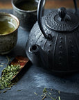 A close-up of a black, ornate cast iron teapot with two small ceramic cups filled with Sencha Kyoto Green Tea by Magic Hour on a dark slate surface. Loose leaf tea leaves and a wooden scoop are scattered around, adding to the rustic, tranquil setting.
