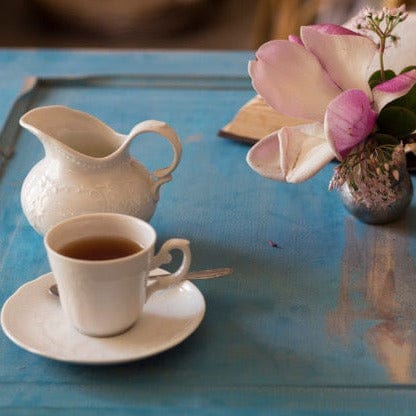 A white teacup filled with Magic Hour Magnolia Oolong Tea sits on a matching saucer alongside a white creamer on a blue table. Next to the tea setup, there is a small vase with pink and white flowers. The background is blurred, focusing on the items on the table.