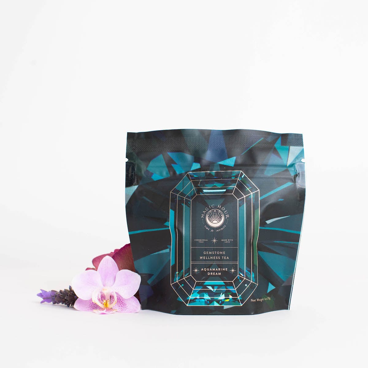 A pouch of &quot;Magic Hour Aquamarine Dream - Soothing Herbal Ayurvedic Adrenal Tonic&quot; stands against a white background. The packaging is designed with a gemstone graphic, featuring teal and black colors. In front of the pouch, there are two delicate pink orchids and a small purple flower, showcasing the allure of this organic loose leaf tea.