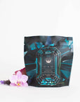 A pouch of "Magic Hour Aquamarine Dream - Soothing Herbal Ayurvedic Adrenal Tonic" stands against a white background. The packaging is designed with a gemstone graphic, featuring teal and black colors. In front of the pouch, there are two delicate pink orchids and a small purple flower, showcasing the allure of this organic loose leaf tea.