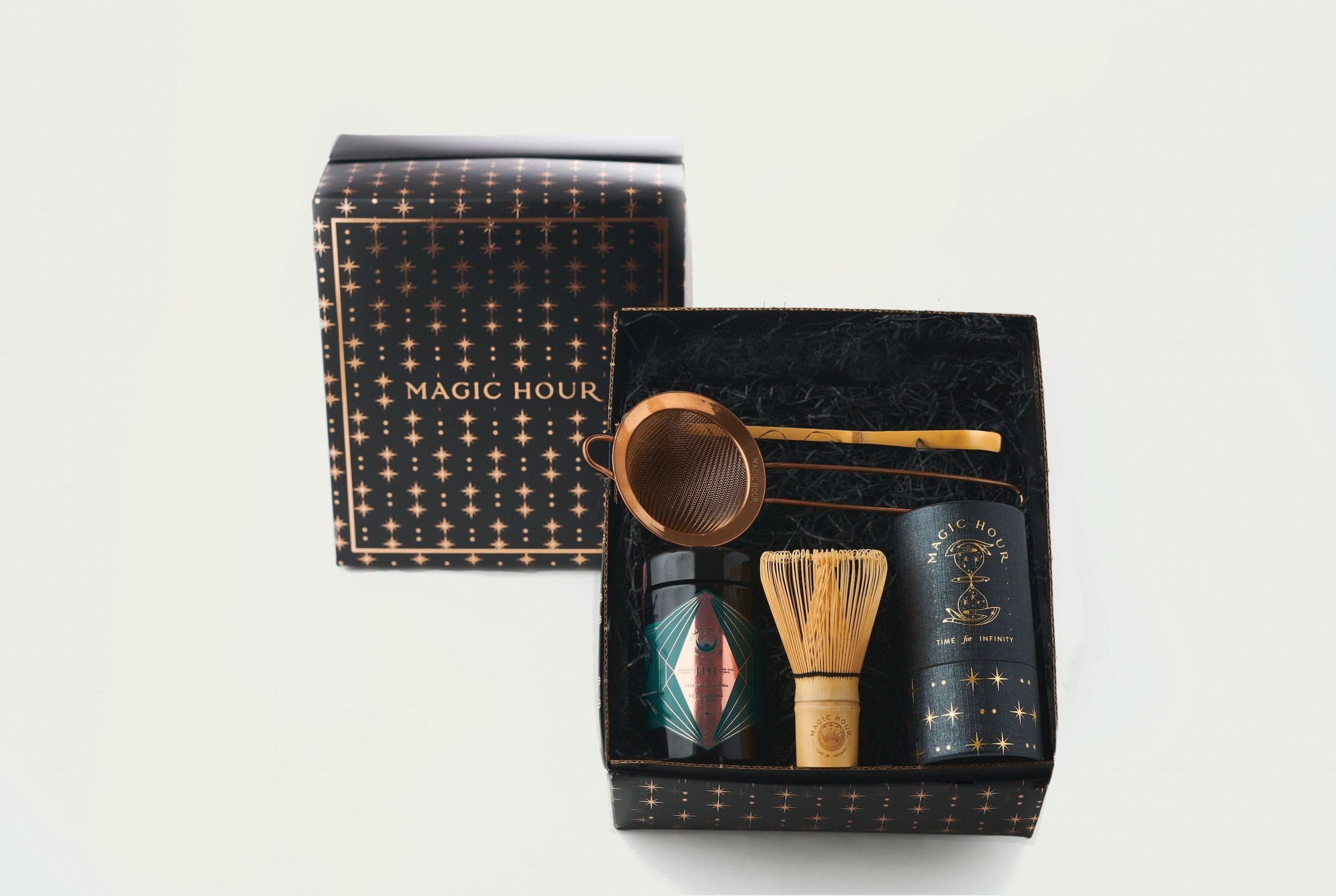A gift box titled "Matcha 1111 : Gift Set" by Magic Hour is open to reveal its contents: a metallic tea strainer, a black tin with a colorful label, organic loose leaf tea, a straw whisk, and a black cylindrical container. The box and lid are black with a pattern of gold stars.