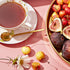 A delicate porcelain teacup from the Flower & Fruit Lovers Starter Set, filled with adaptogenic tea and accompanied by a gold spoon, sits on a pink surface. Nearby, a golden tray holds an assortment of fresh fruits, including figs and cherries. The scene is beautifully accented with a white flower and additional cherries scattered around.