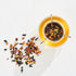 A top-down view of The Astrology Collection - Luxe Sampler Sips glass teapot filled with brewed rare teas. A metal infuser with dried tea leaves sits inside the teapot. Loose tea leaves are scattered beside the teapot on a white surface. Sunlight casts a soft shadow, adding a warm, cozy ambiance.