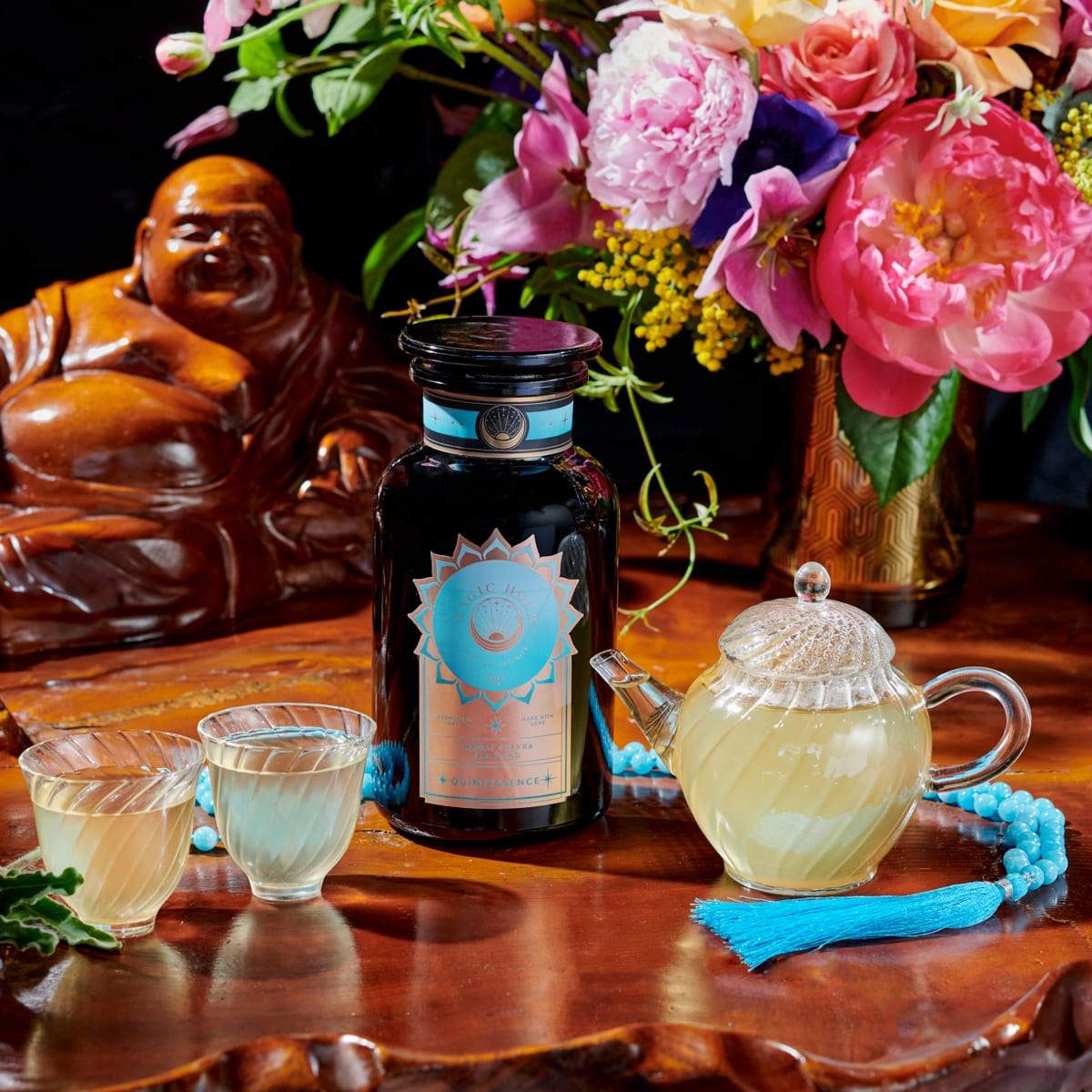 A dark bottle with an ornate label is placed on a wooden surface beside two glass cups, a clear teapot filled with light yellow organic tea, a strand of blue beads, and a colorful bouquet of flowers. A statue is visible in the background. The label reads "Quintessence™ Tea for Opening & Healing the Throat Chakra" by Magic Hour.