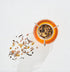 A glass cup of Classics - Luxe Sampler Sips tea rests on a white surface, adorned with a metal tea strainer on top. Loose tea leaves and spices are scattered around, their shadows softly cast in the background light. The amber hue of the tea stands out against the pristine, white backdrop.