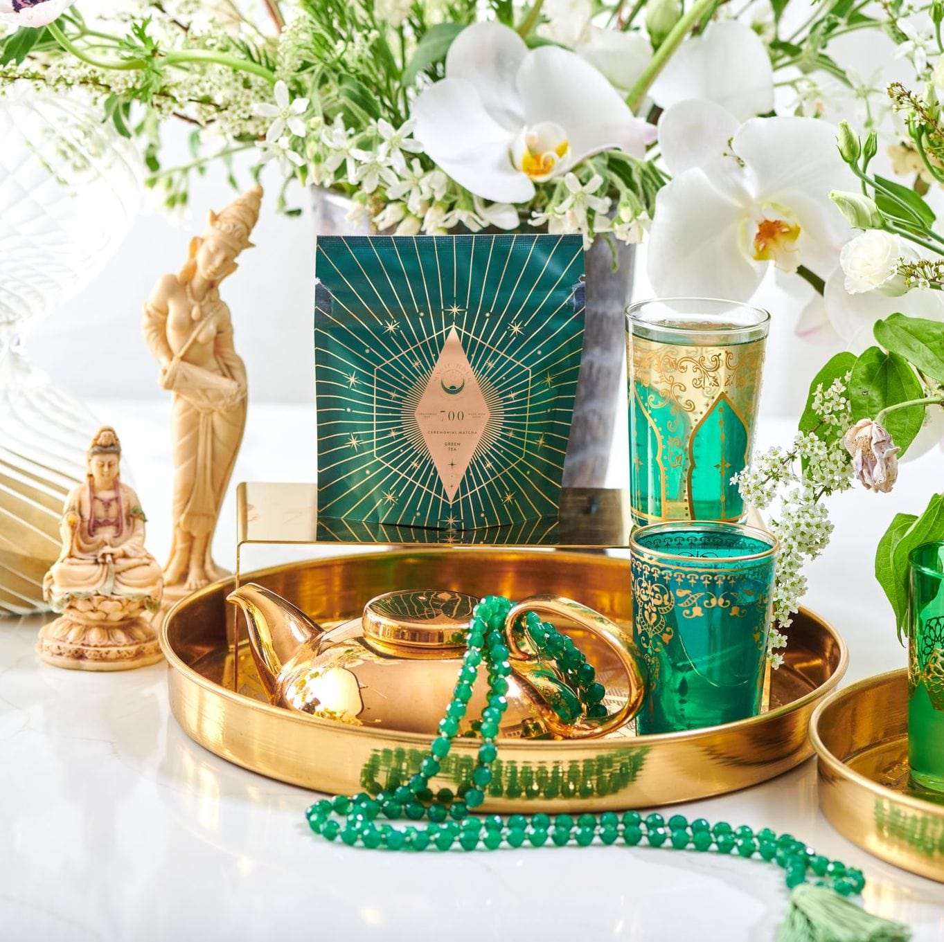 A decorative setup featuring a gold tray with a teapot, green beaded necklace, and ornate green cups. In the background are white flowers, greenery, a bag of Club Magic Hour Organic Ceremonial Matcha 700, and two statuettes of women. The organic tea enhances the fresh ambiance.