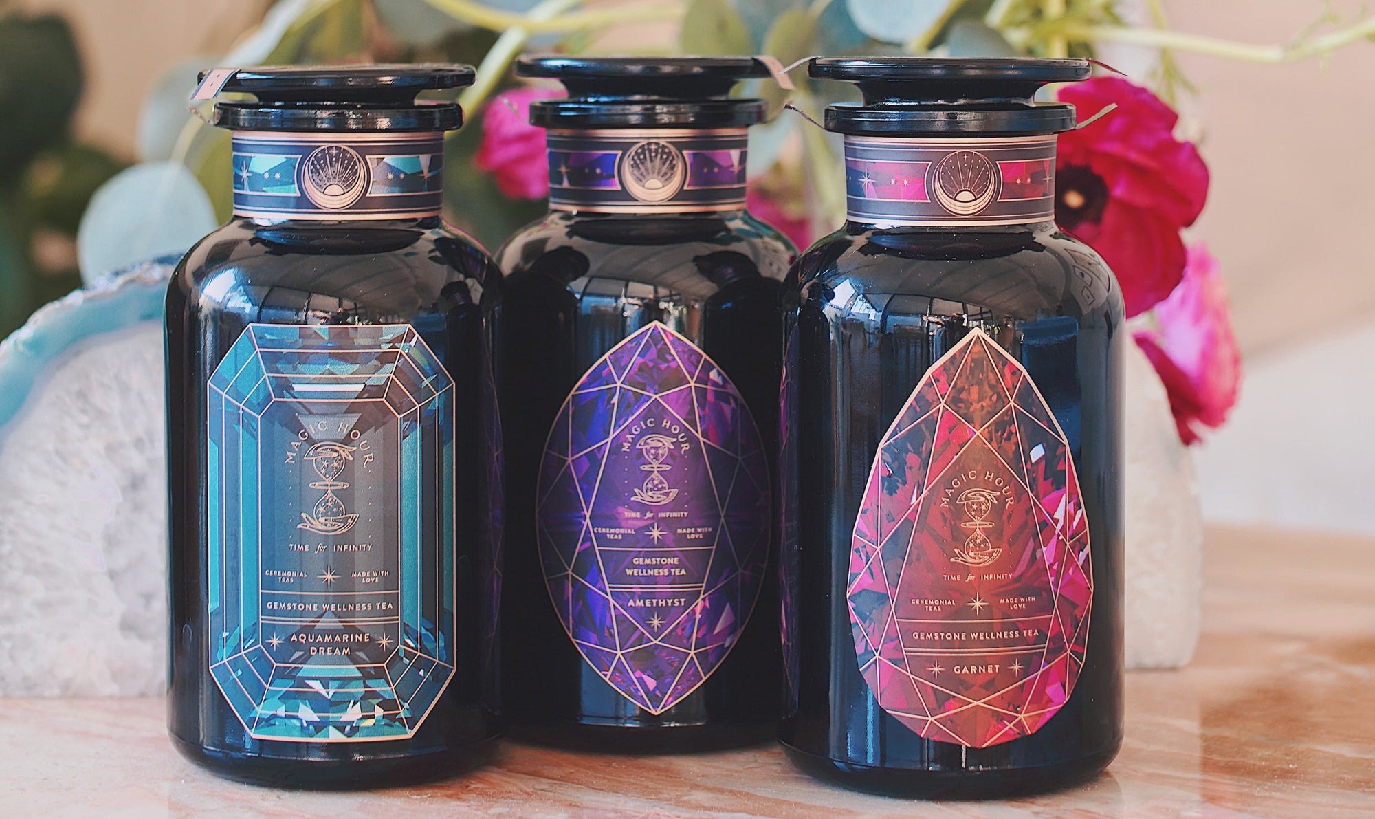 Three dark glass bottles with ornate, geometric labels are displayed side by side on a wooden surface. The labels feature intricate crystal designs in blue, purple, and red tones. A blurred background includes green leaves and a pink flower, enhancing the allure of the **Magic Hour Gemstone Bundle: Garnet, Amethyst, Aquamarine**.