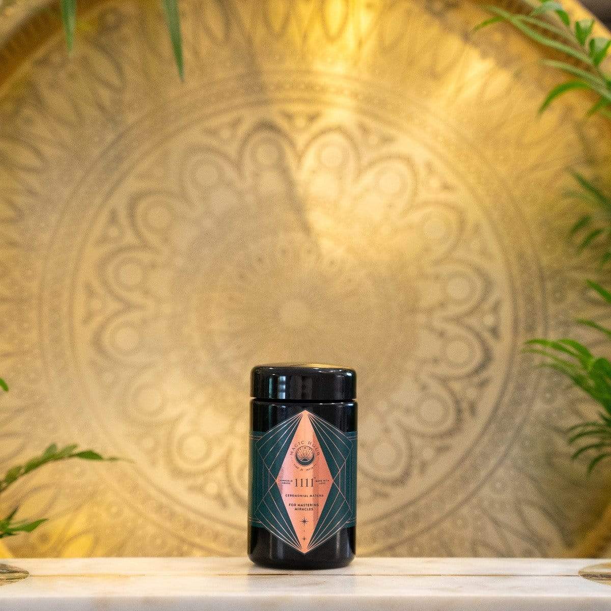 A black cylindrical container labeled with geometric pink and green designs rests on a white surface. In the background, an ornate, circular golden pattern adorns the wall, framed by green plants on either side. This serene and elegant setting perfectly complements your Matcha 1111 : Gift Set experience by Magic Hour.


