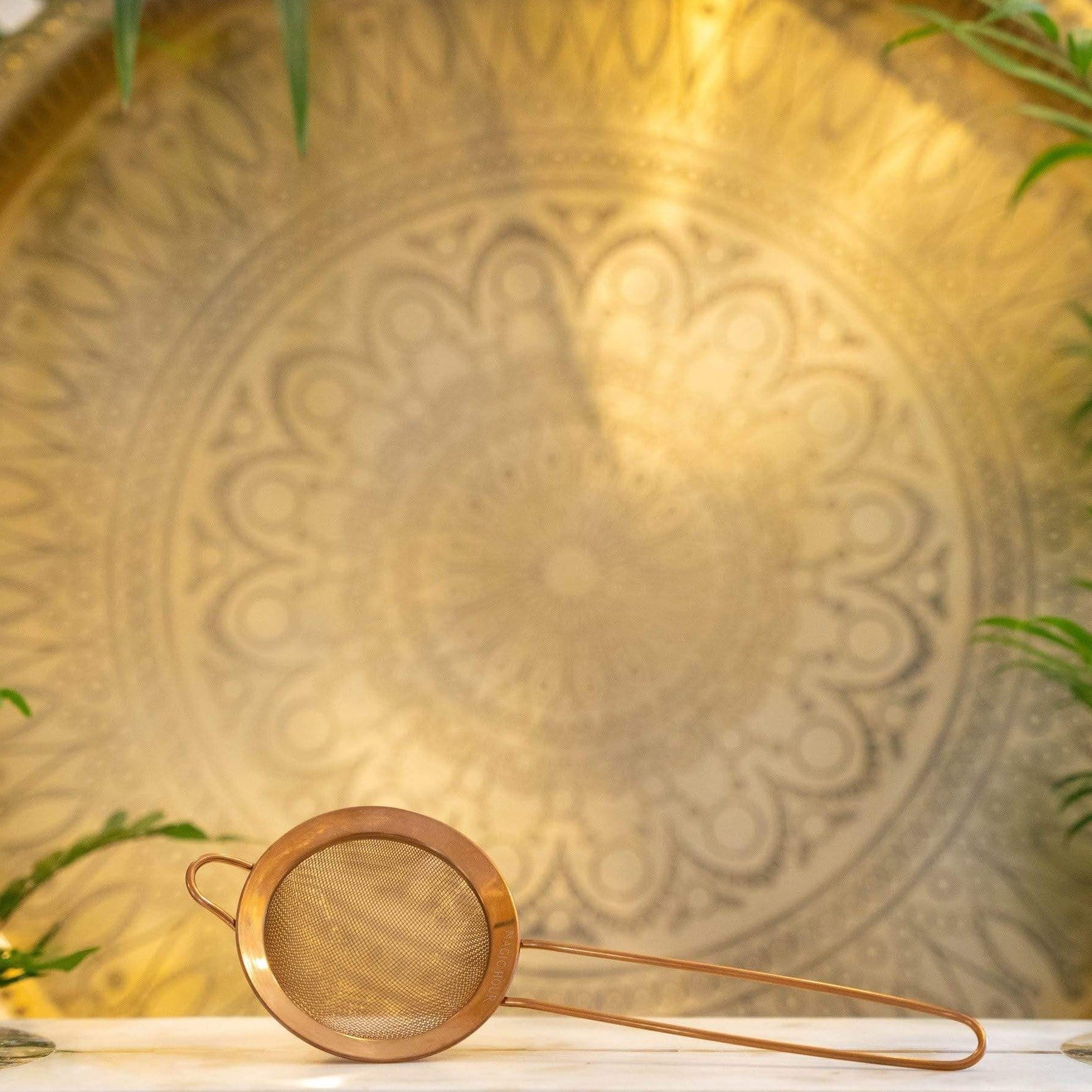 A Rose Gold Matcha Sift rests on a white surface with a detailed, golden mandala pattern in the background, illuminated by warm, soft lighting. Green foliage partially frames the scene, contributing to the serene and elegant ambiance—perfect for a moment with Magic Hour.