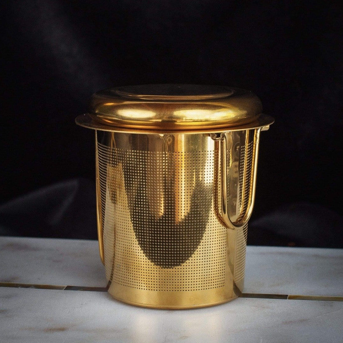 A gold-toned metallic tea infuser with a cylindrical shape and a perforated body, featuring a fitted lid and a handle, is perfect for brewing your favorite Loose Leaf Tea. The PMB Mesh Screen Company's Midas Touch: Golden-Hued Tea Strainer is placed against a dark background on a light-colored surface.