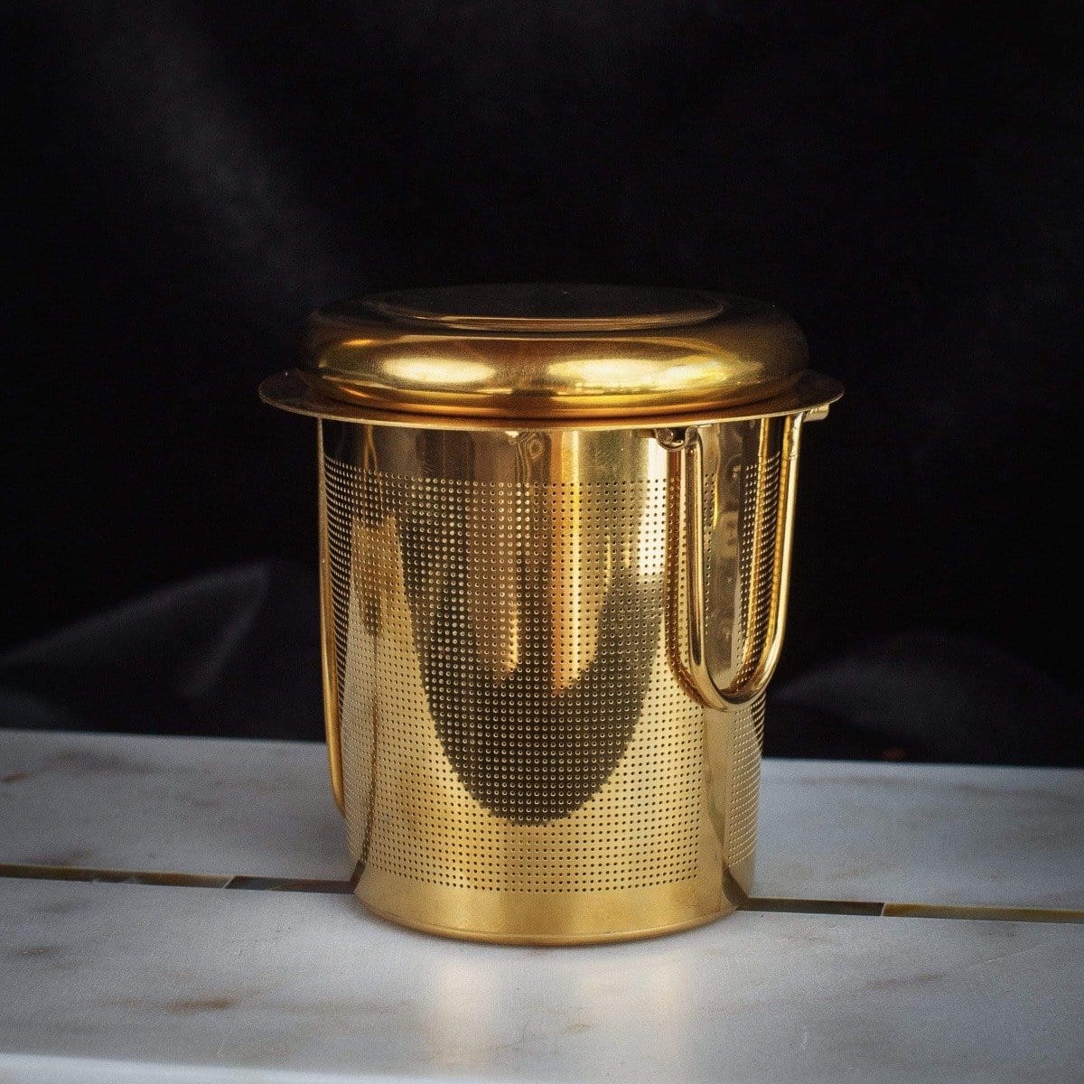 A gold-toned metallic tea infuser with a cylindrical shape and a perforated body, featuring a fitted lid and a handle, is perfect for brewing your favorite Loose Leaf Tea. The PMB Mesh Screen Company's Midas Touch: Golden-Hued Tea Strainer is placed against a dark background on a light-colored surface.