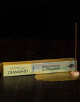 A horizontal box of Diamond Incense from Shoyeido rests on a dark wooden surface. To the right of the box, an incense stick rich with sandalwood and other fragrances is lit and held upright in a small holder, with a delicate golden mesh beneath it.
