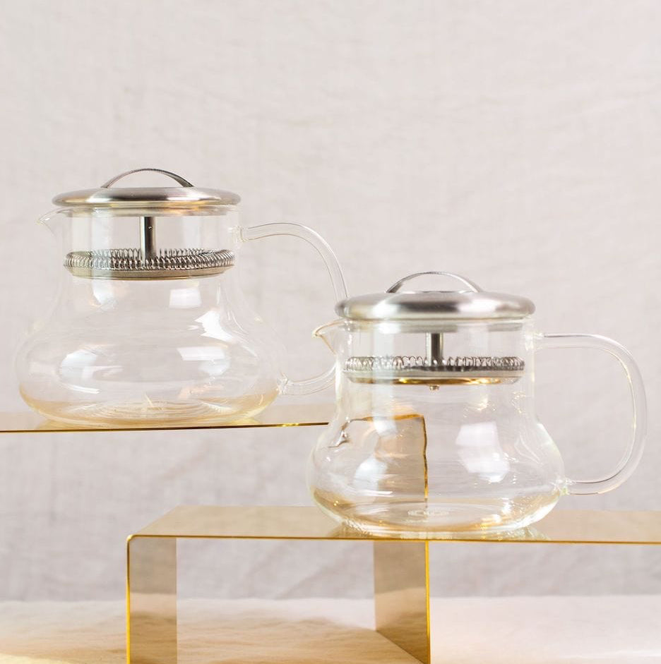 Two Essential Perfection Teapots with Built-in Strainers and metal lids are displayed on two yellow rectangular stands of different heights. These teapots, featuring built-in strainers, have a simple, modern design with round bodies, curved handles, and small spouts—perfect for enjoying loose leaf tea against the plain white fabric background.