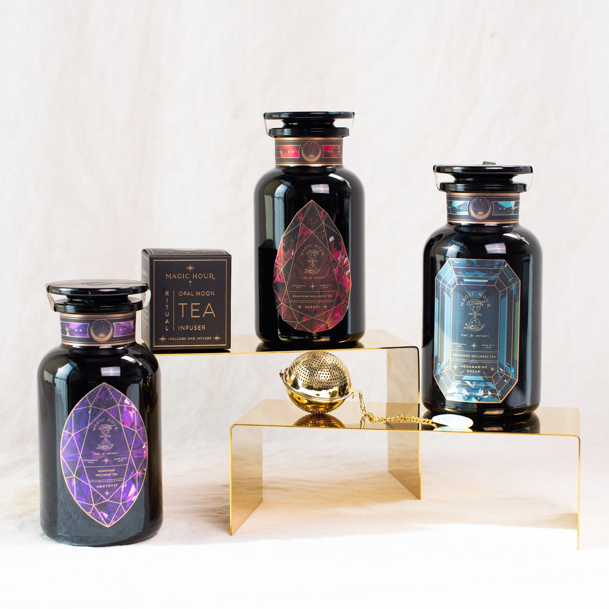 The image shows three black glass jars of Magic Hour Gemstone Bundle: Garnet, Amethyst, Aquamarine with colorful geometric labels, arranged on golden risers. Each label features intricate designs in different colors: purple, red, and blue. A small black box labeled "Magic Hour Tea Infuser" and a gold tea infuser are also displayed.