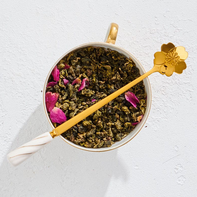 A cup filled with loose-leaf tea, adorned with pink flower petals, features the elegant Gold Floral Teaspoon resting across the top. The teaspoon, with its long gold stem and delicate white handle ending in a flower shape, is placed on a white textured surface.