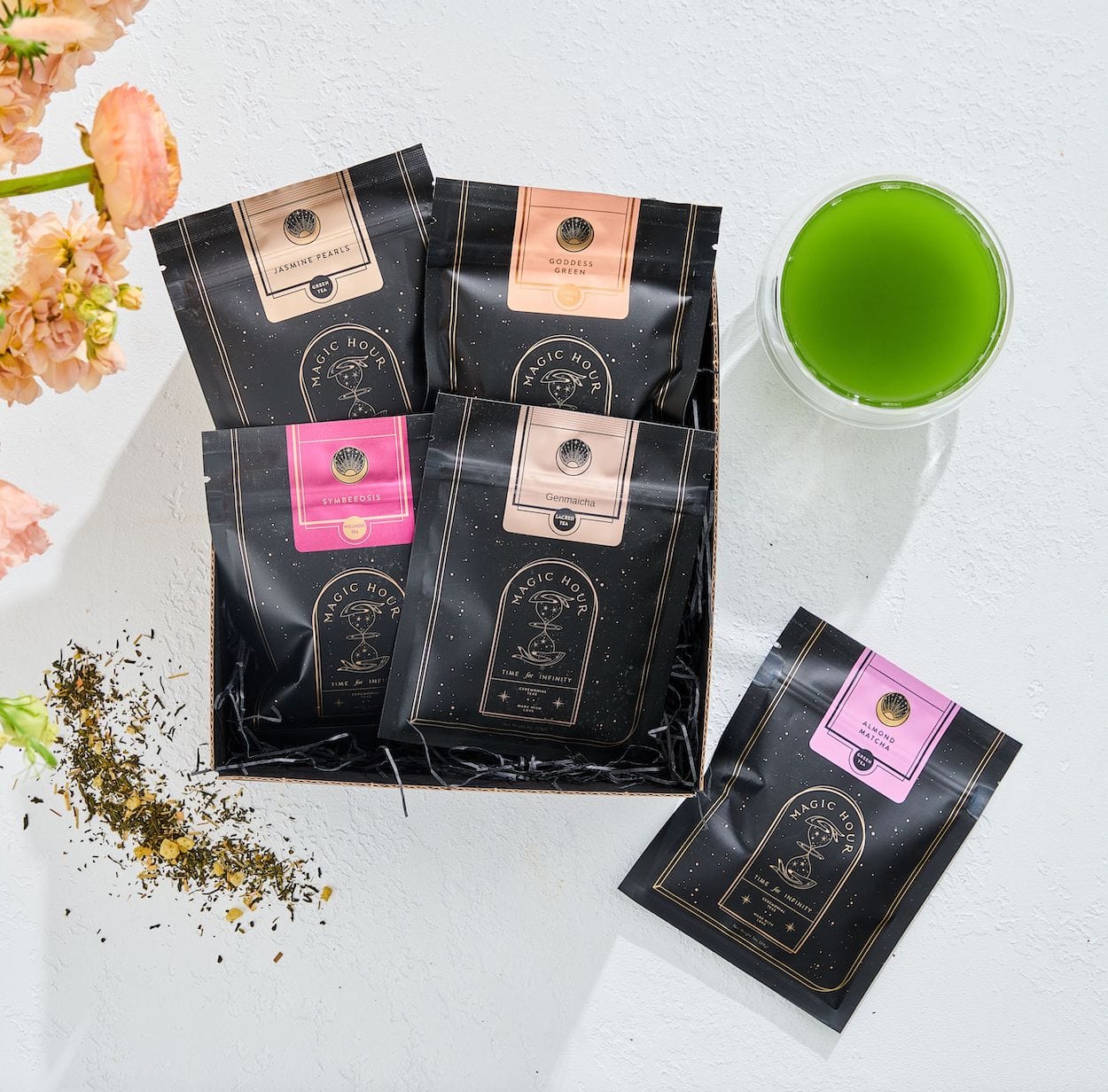 A display of five black packets of Green Magic Sampler Box from "Magic Hour" in various flavors, laid out on a white surface alongside a glass of vibrant green tea. Some loose leaf tea is scattered near the packets, and a bouquet of peach-colored flowers is partially visible.