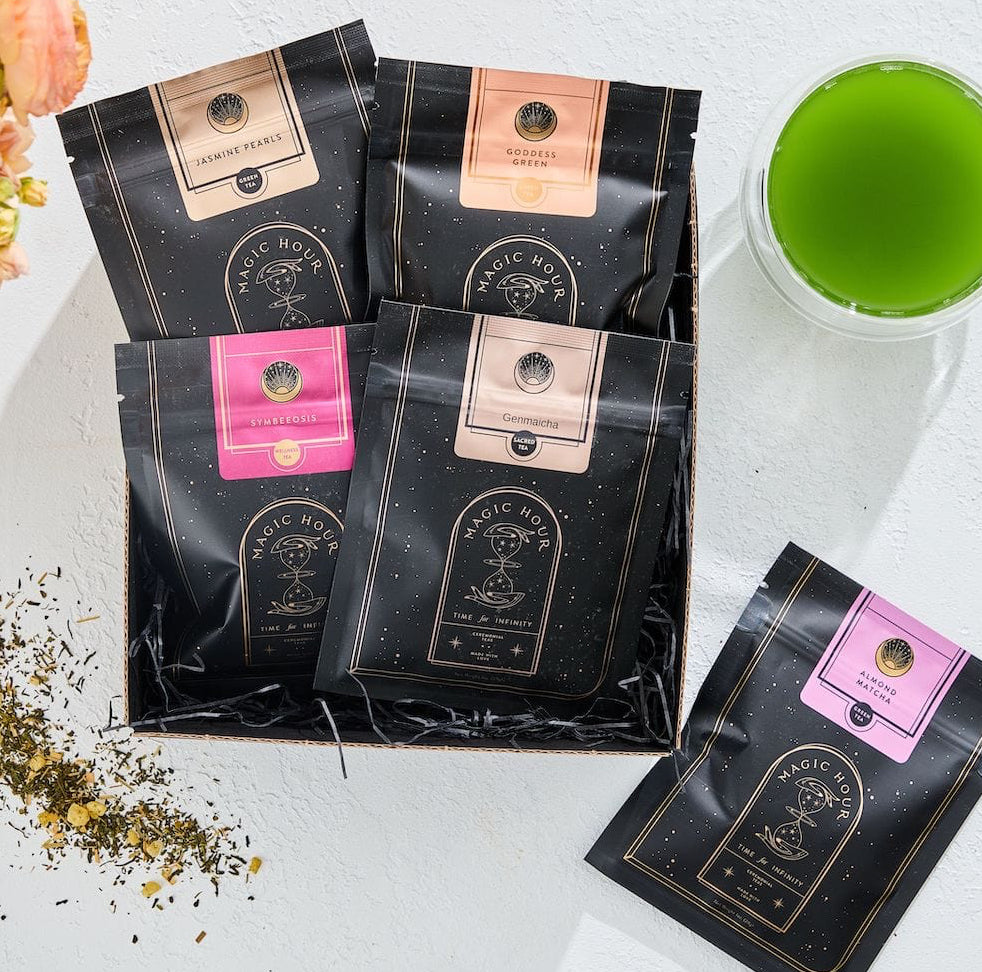A display of five black packets of Green Magic Sampler Box from "Magic Hour" in various flavors, laid out on a white surface alongside a glass of vibrant green tea. Some loose leaf tea is scattered near the packets, and a bouquet of peach-colored flowers is partially visible.