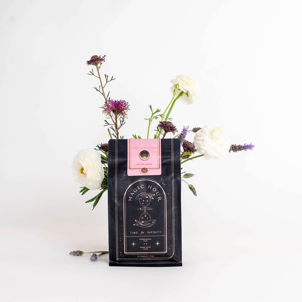 A black and pink package of "Gypsy Rose Black Tea" by Magic Hour stands on a white surface, adorned with the label "Time for Infinity". A few white and purple flowers, including lavender, are creatively arranged around and behind the package, giving it a fresh, elegant look.