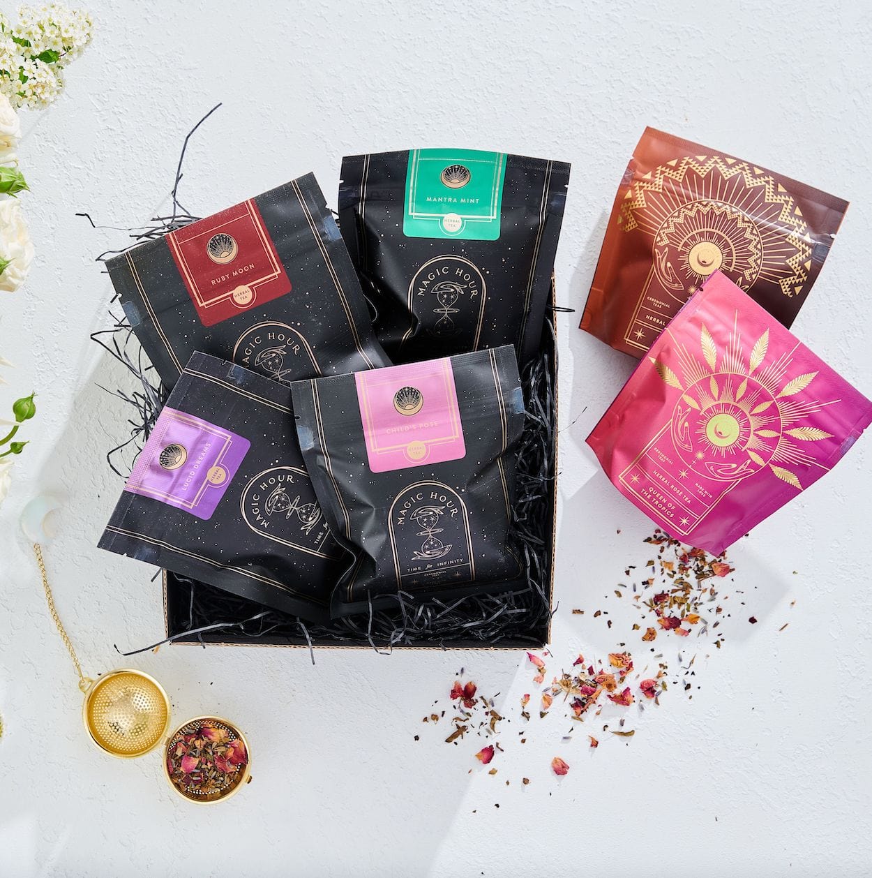 A box filled with assorted packages of loose leaf tea is displayed. The packages showcase various vibrant designs and colors, including black, pink, red, and green. A gold tea infuser with dried organic tea leaves is placed beside the **Magic Hour Herbal Magic Sampler Tea Box: Caffeine-Free Teas for Sleep, Digestion & Immunity** on a white surface.