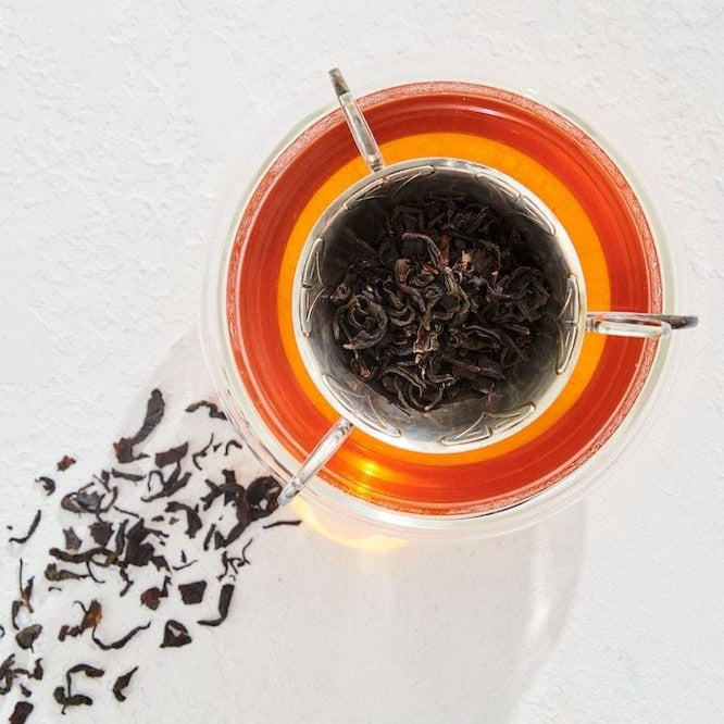 A glass cup filled with amber-colored Tea of The Rising Sun: Japanese Breakfast from Magic Hour sits on a white surface, with a metal tea infuser containing loose black tea leaves inside the cup. Loose leaf tea is scattered next to the cup, casting a shadow on the surface.