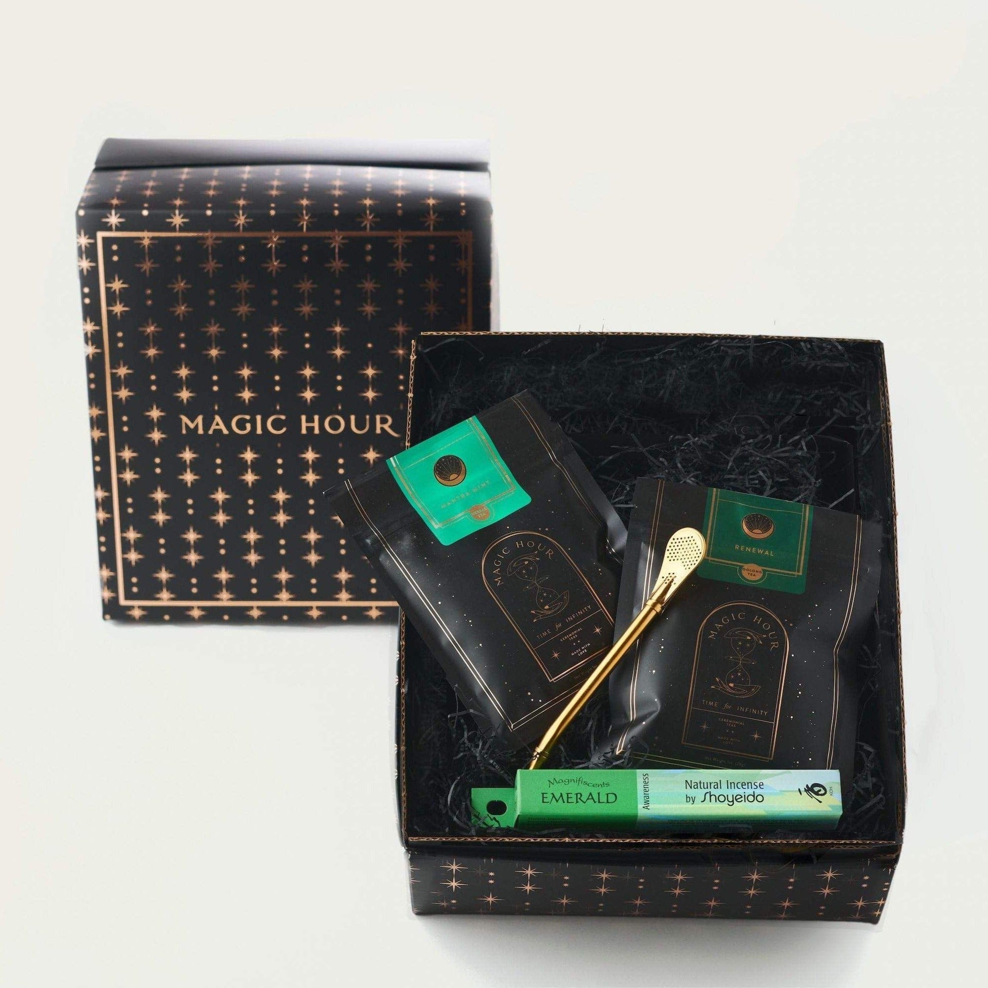 A black box labeled "Renew & Relax: Ritual Gift for Mindfulness & Calm by Magic Hour" is open, revealing its contents. Inside are two black packages with green labels and a gold metallic spoon. Another green box labeled "Emerald Natural Incense by Shoyeido" lies beside the packages inside the box, creating a serene setting for your organic tea experience.