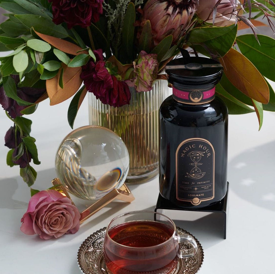 A colorful floral arrangement sits in a vase on a table next to a bottle labeled "Magic Hour" and a teacup filled with Soulmate: Chocolate-Raspberry-Rose Black Tea for Finding & Celebrating Love by Magic Hour. A glass orb and a single pink rose are also on the table, creating an elegant and inviting scene.