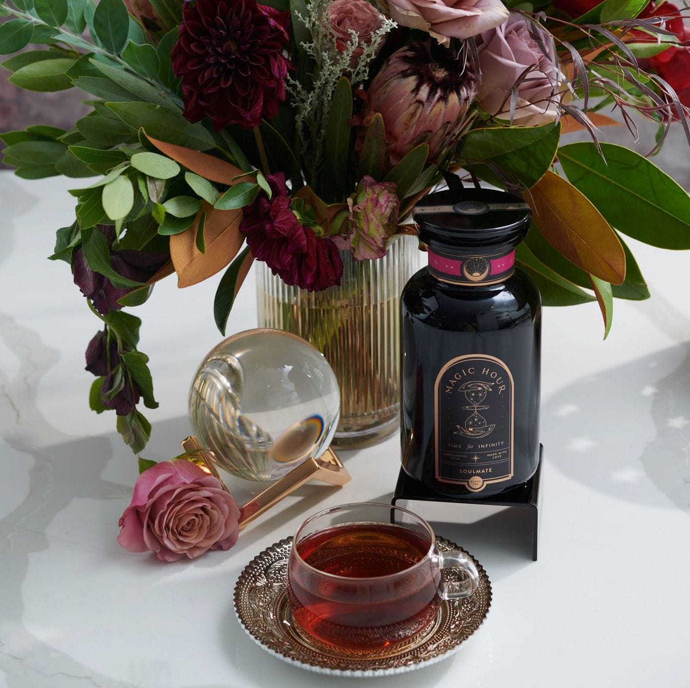 A colorful floral arrangement sits in a vase on a table next to a bottle labeled &quot;Magic Hour&quot; and a teacup filled with Soulmate: Chocolate-Raspberry-Rose Black Tea for Finding &amp; Celebrating Love by Magic Hour. A glass orb and a single pink rose are also on the table, creating an elegant and inviting scene.