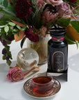 A colorful floral arrangement sits in a vase on a table next to a bottle labeled "Magic Hour" and a teacup filled with Soulmate: Chocolate-Raspberry-Rose Black Tea for Finding & Celebrating Love by Magic Hour. A glass orb and a single pink rose are also on the table, creating an elegant and inviting scene.