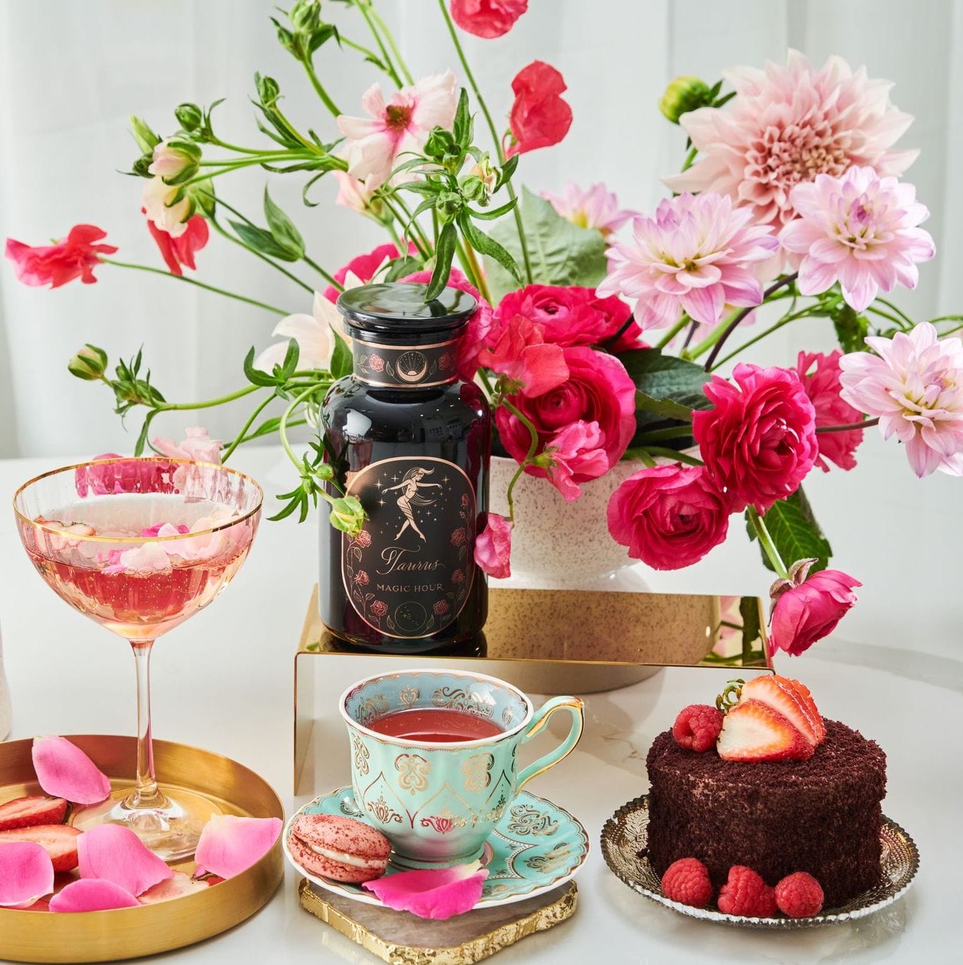 A beautifully arranged table featuring a bouquet of pink and red flowers, a glass of pink beverage with rose petals, a turquoise teacup filled with Magic Hour Taurus: Tea of Venusian Garden Delights and its matching saucer, a pink macaron, a small chocolate cake garnished with strawberries and raspberries, and a bottle labeled "Glamour Magic Rocks.