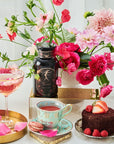 A beautifully arranged table featuring a bouquet of pink and red flowers, a glass of pink beverage with rose petals, a turquoise teacup filled with Magic Hour Taurus: Tea of Venusian Garden Delights and its matching saucer, a pink macaron, a small chocolate cake garnished with strawberries and raspberries, and a bottle labeled "Glamour Magic Rocks.