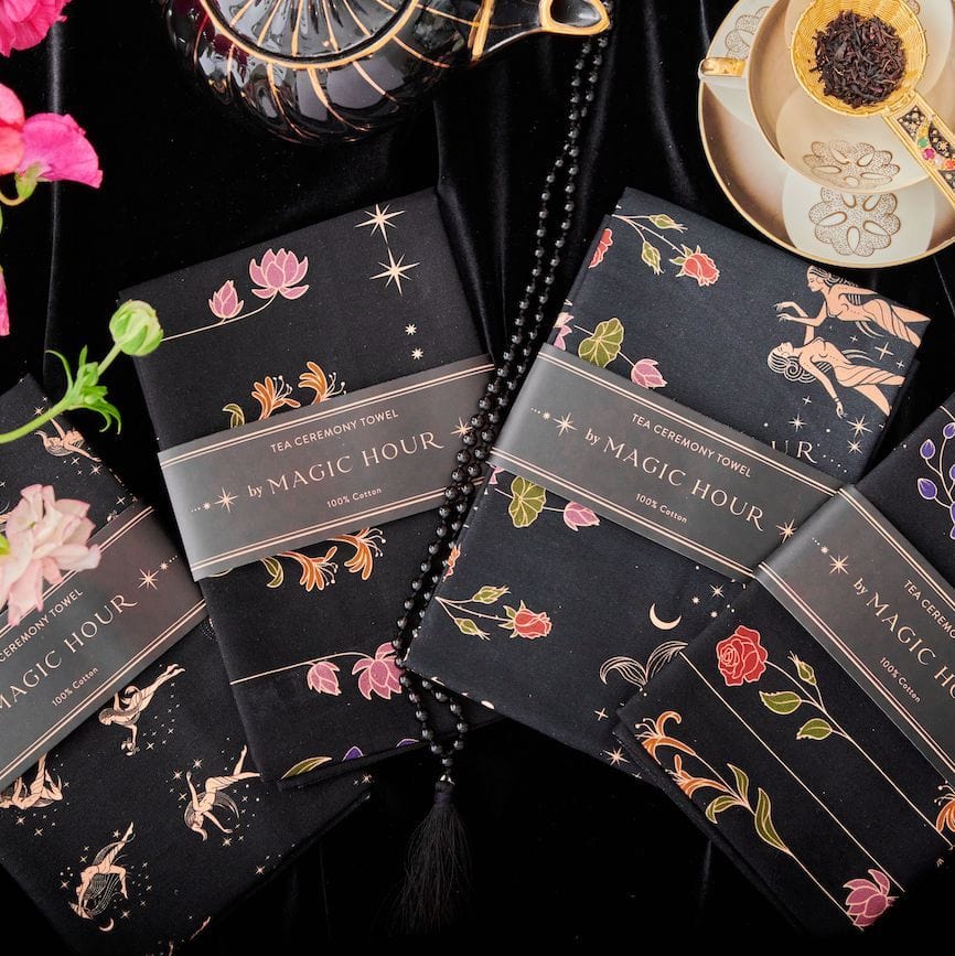 A display of folded Tea Ceremony Towels featuring various floral and botanical designs on a black background. The towels are branded "Magic Hour" and are arranged near a decorative teapot, a string of beads, flowers, and a tea cup with a tea strainer, perfect for enjoying your organic tea.