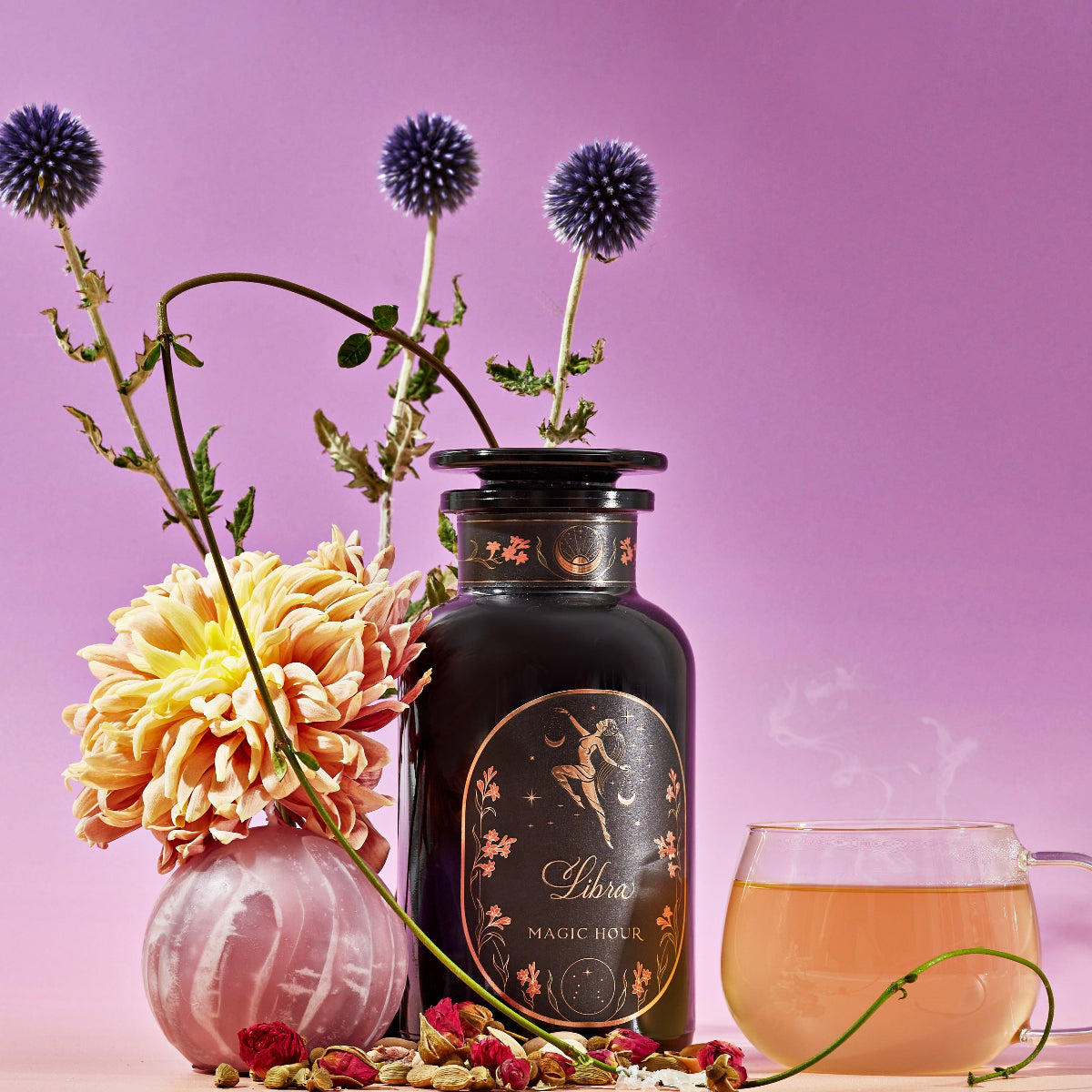A pink and purple-toned still life image featuring a black bottle labeled "Magic Hour," adorned with intricate gold designs. Surrounding it are vibrant flowers, a glass teacup with steaming Libra- Pistachio-Rose Persian Love Cake Tea with White Pearls & Shatavari, dried herbs, and purple thistle-like flowers.
