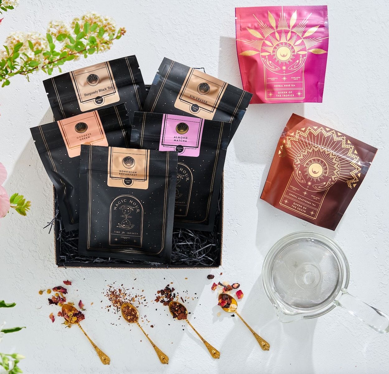 A display of various packaged tea blends, including Magic Hour's The Best of Magic Hour Sampler Set with Tea in Hand Teapot, features five black and two colored packets. The black packets have copper labels. Beside the packages are three gold spoons with loose leaf tea, a clear glass teapot, and green foliage providing a decorative touch.