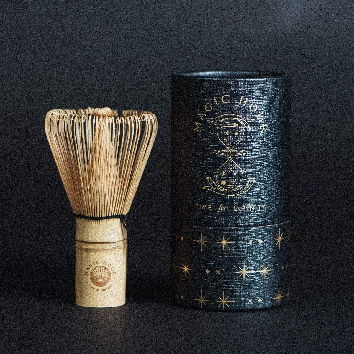 An Artisanal Magic Hour Matcha Whisk designed for matcha tea preparation stands next to a cylindrical black container adorned with gold celestial designs and the text "MAGIC HOUR" and "TIME for INFINITY." Both items, perfect accompaniments to your organic tea ritual, are set against a dark background.