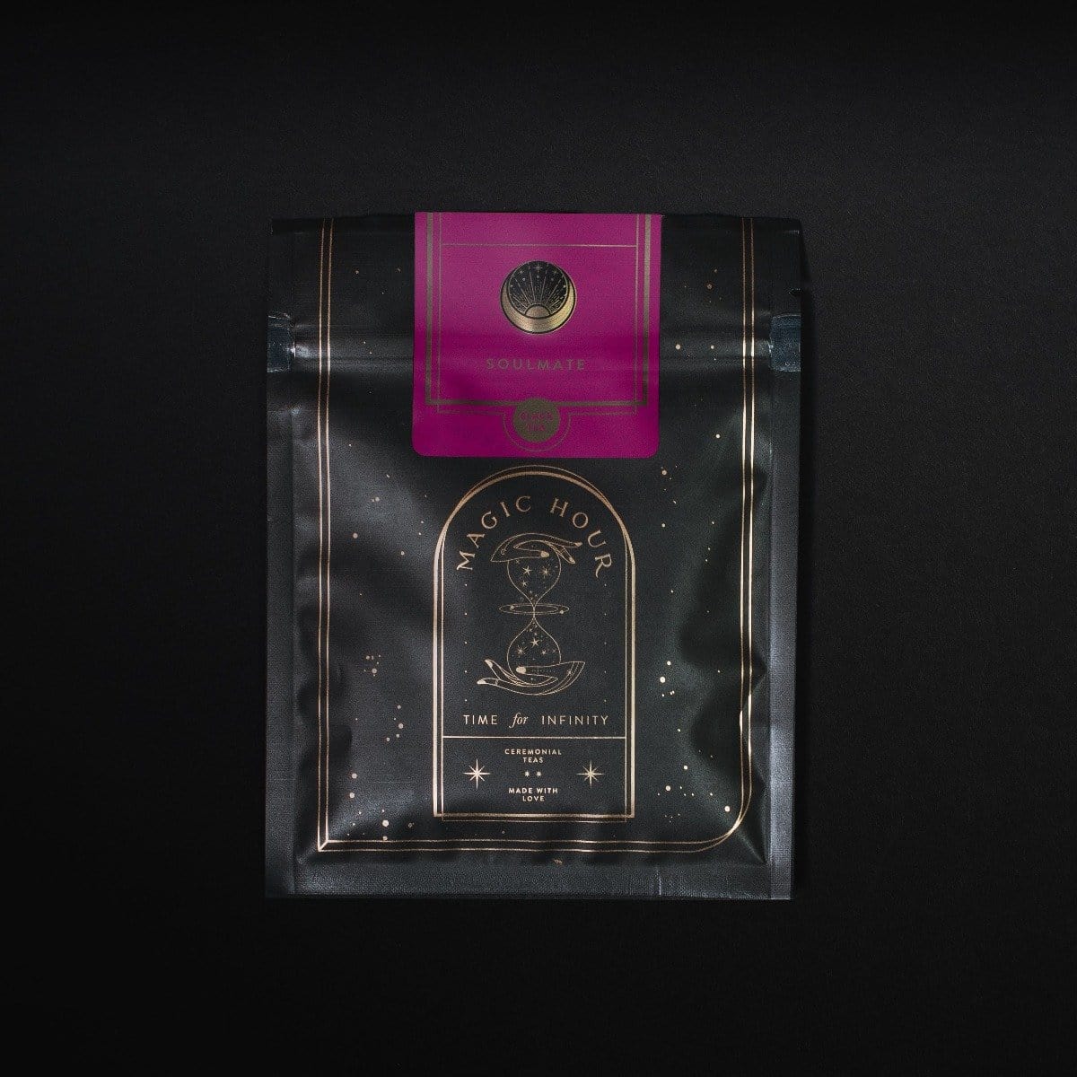 A black-colored Magic Hour Tea package is shown against a dark background. The bag has a decorative gold design with an hourglass illustration, and it reads &quot;Time for Infinity.&quot; A pink label with a circular emblem that says &quot;Soulmate: Chocolate-Raspberry-Rose Black Tea for Finding &amp; Celebrating Love&quot; is attached at the top, highlighting its organic loose leaf tea inside.