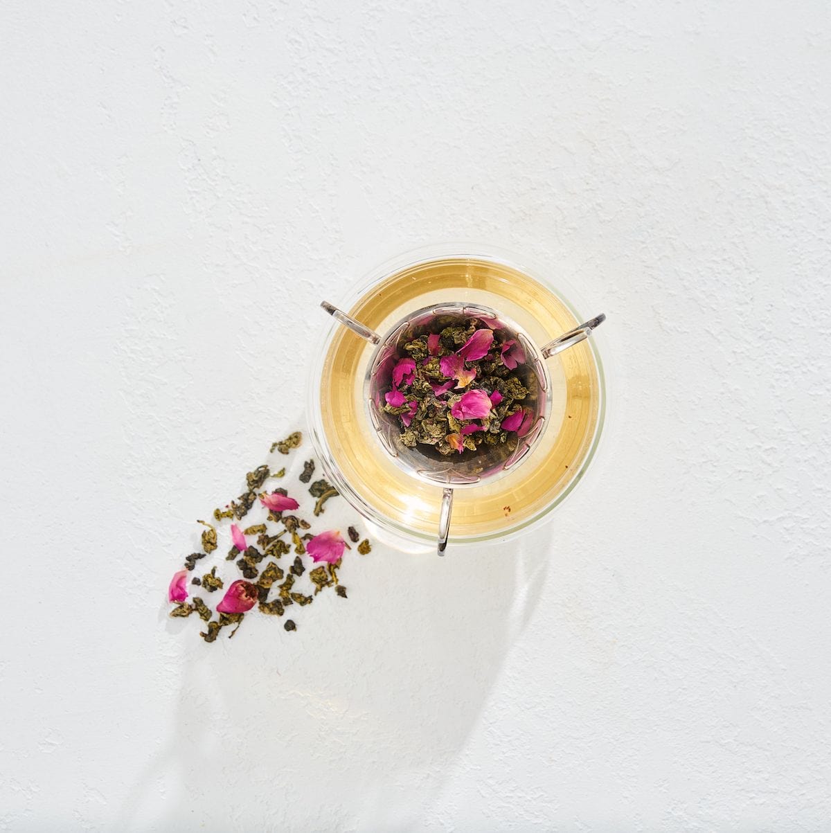 A glass teacup filled with light-colored tea and rose petals sits on a white surface. Loose leaf Magnolia Rose Oolong Tea and rose petals are scattered next to the cup, evoking the serene ambiance of a Magic Hour ritual.