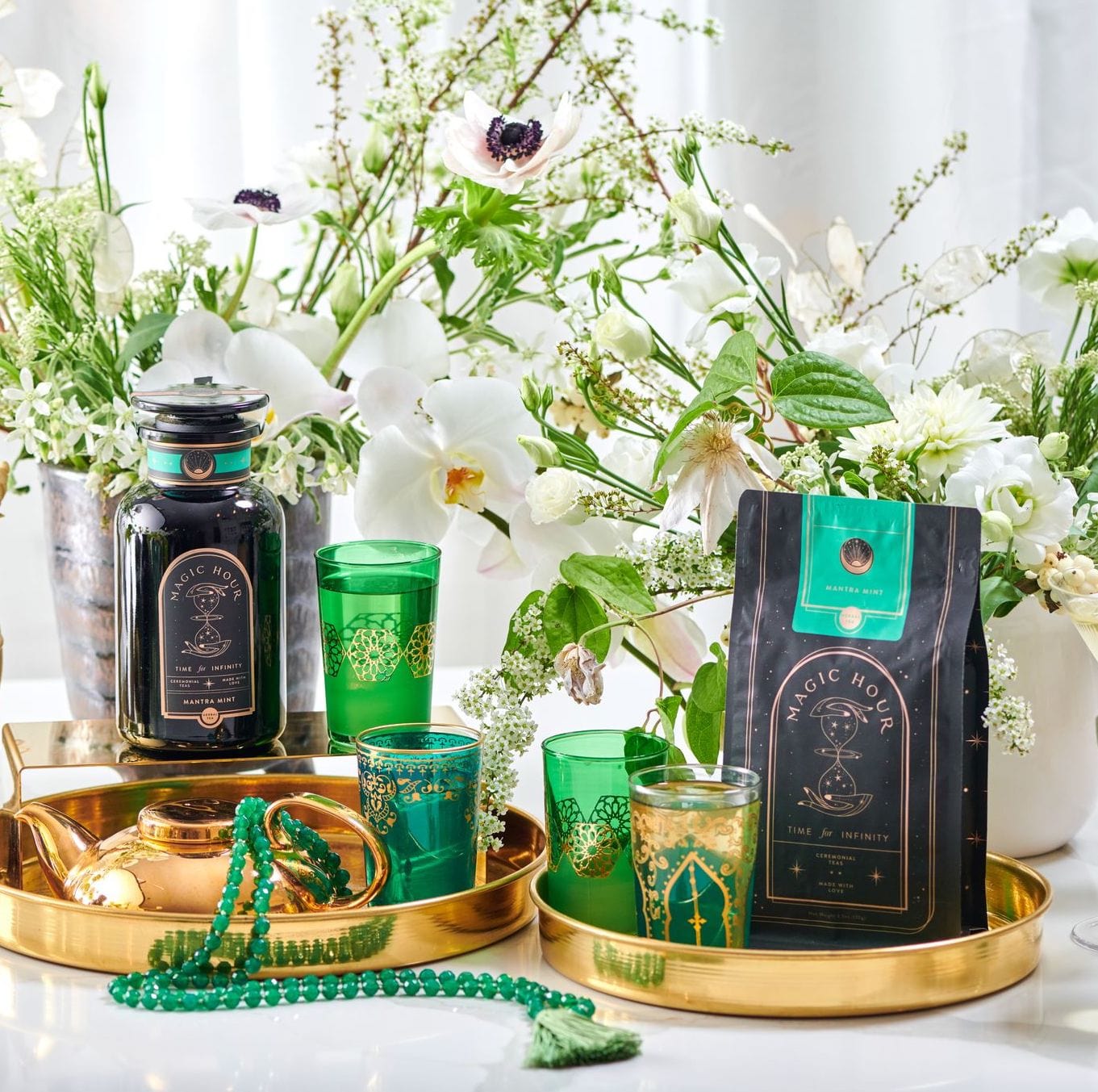 A luxurious tea setup features a black canister of Mantra Mint™ Herbal Tea from Club Magic Hour and a matching bag displayed on a golden tray. Green glasses adorned with golden patterns, a teapot, and a green beaded necklace complement the arrangement, surrounded by white flowers.
