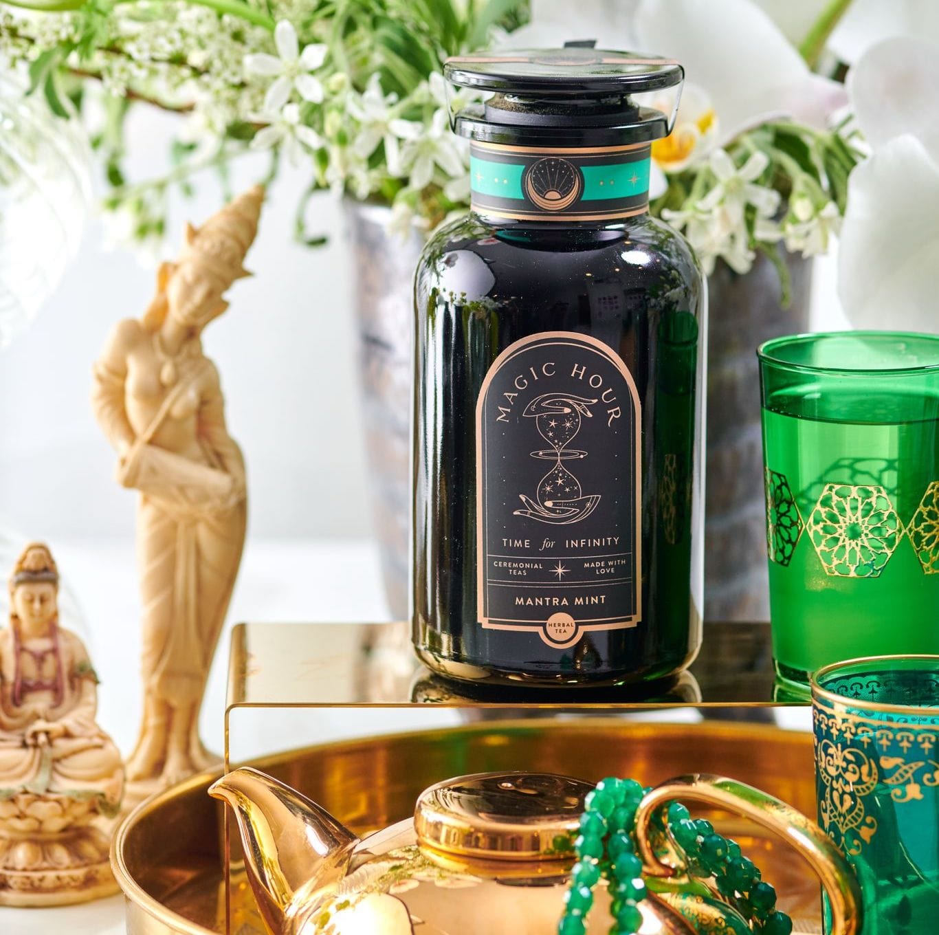 A teal and gold-themed tea setup featuring a black jar of "Mantra Mint™ Herbal Tea" by Club Magic Hour with an intricate label, surrounded by vibrant green glasses, a golden teapot, beads, and statues. It's set on a reflective golden surface with flowers in the background.