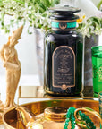A teal and gold-themed tea setup featuring a black jar of "Mantra Mint™ Herbal Tea" by Club Magic Hour with an intricate label, surrounded by vibrant green glasses, a golden teapot, beads, and statues. It's set on a reflective golden surface with flowers in the background.