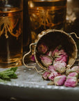 Close-up of a tea setup featuring a decorative bronze tea strainer with dried pink rosebuds spilling out onto a white tray. Two ornate glasses with golden accents contain Mantra Mint™ Herbal Tea by Club Magic Hour, a light amber organic tea, and fresh green mint leaves are placed on the tray.