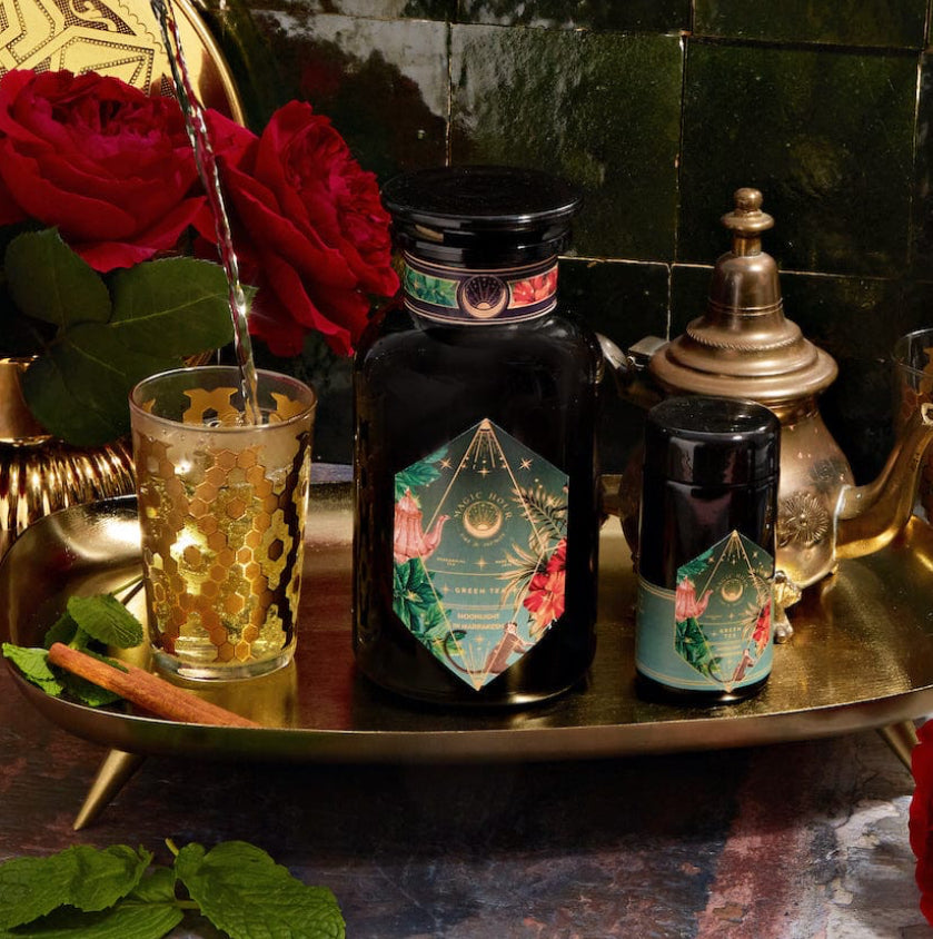 A decorative scene featuring a dark bottle with intricate golden and colorful designs, accompanied by a matching smaller container on an ornate golden tray showcases the opulence of Moonlight in Marrakesh: Spiced Mint Green Tea Case Packs by Magic Hour. Two lavishly adorned glasses are being filled with this exquisite Moroccan mint tea, while red roses and a teapot accentuate the setting.