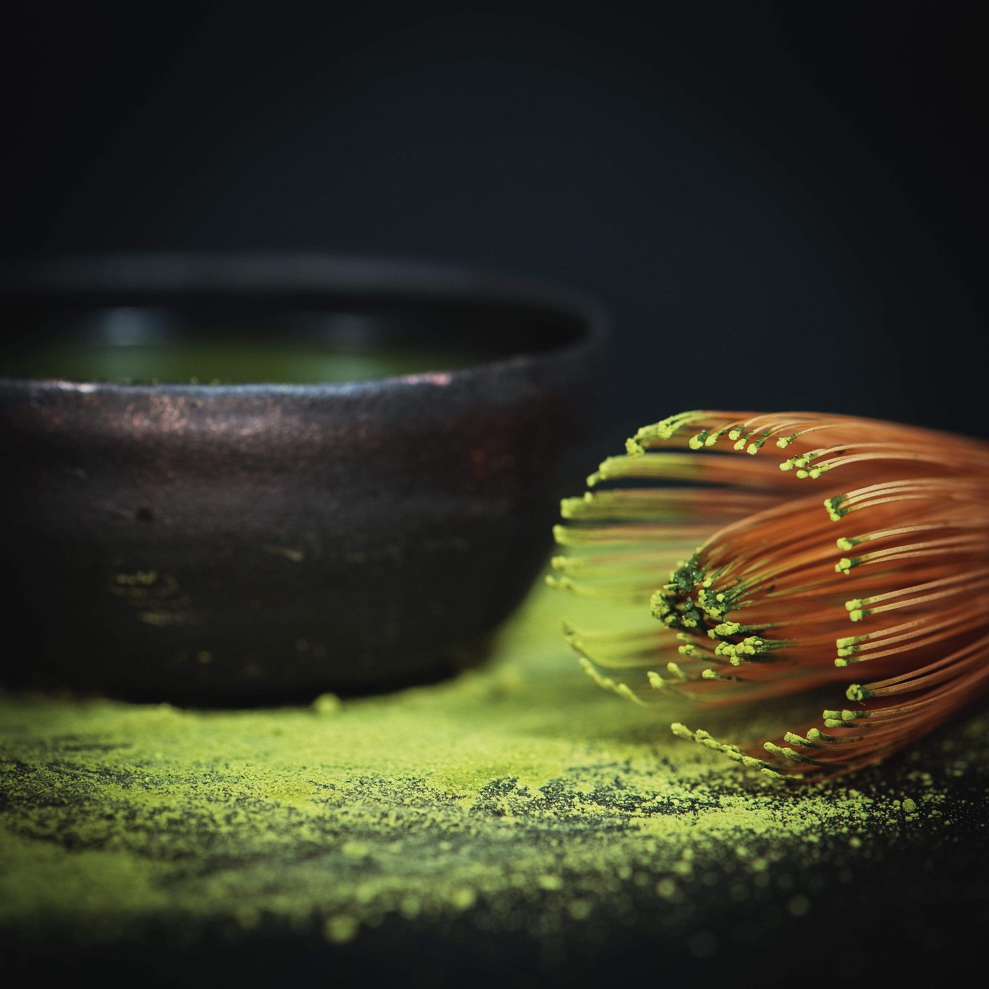 A Ceremonial Matcha Freedom Box with green matcha powder on its bristles lies next to a rustic brown ceramic bowl. The surface is speckled with organic tea powder from Magic Hour, creating a vibrant contrast against the dark background.
