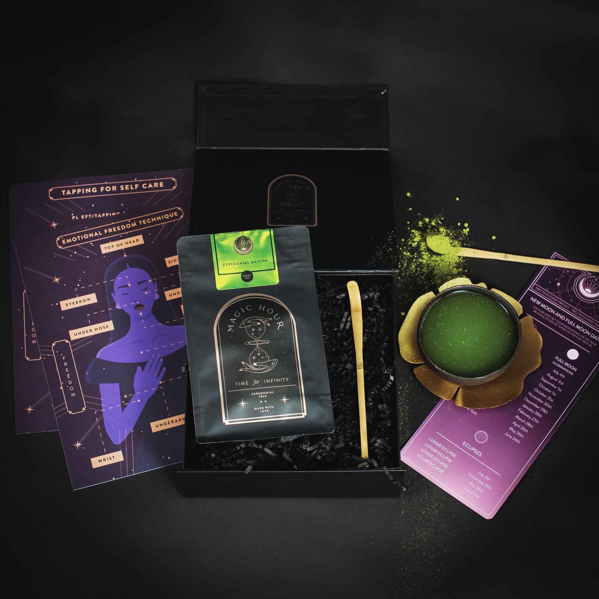A flat lay of a black box containing a tea package labeled "Ceremonial Matcha Freedom Box by Magic Hour," a ceramic cup with green tea powder, a gold spoon, and informational pamphlets. The items are arranged artfully on a black background with loose leaf tea and green tea powder scattered around.