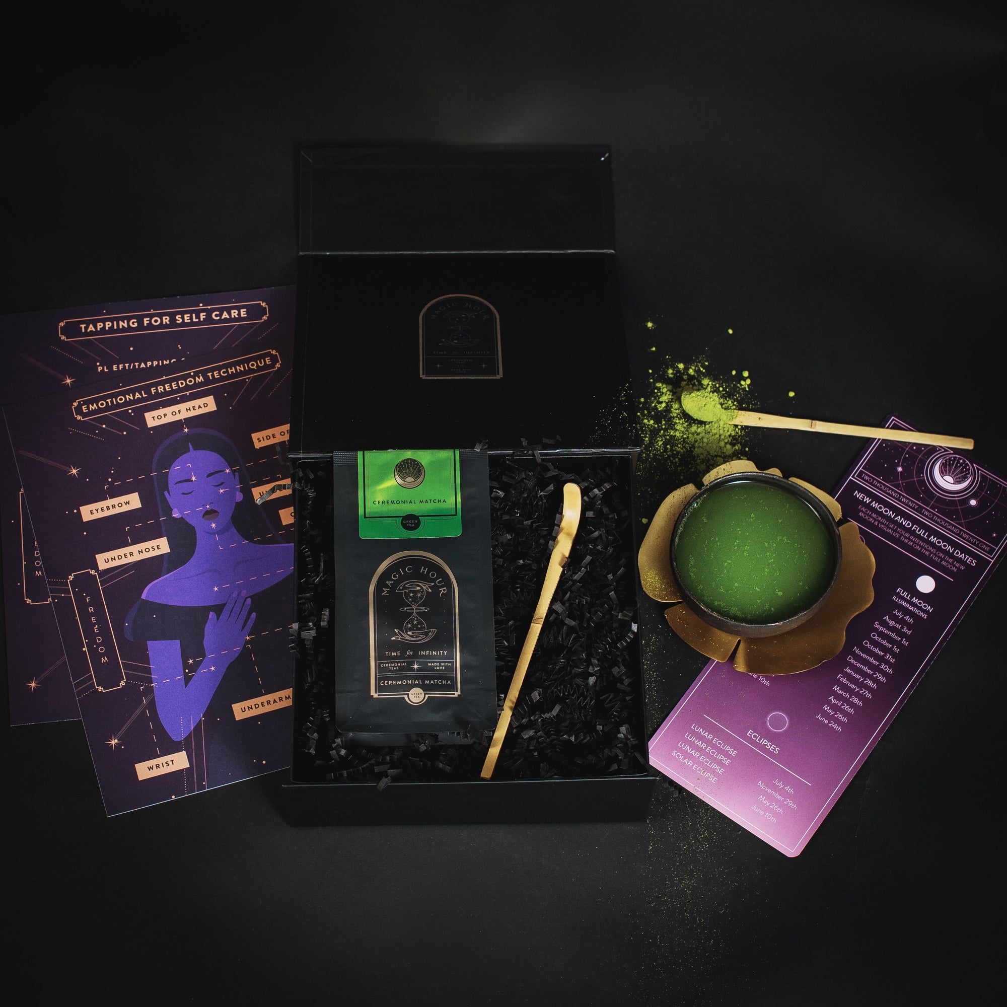 A self-care kit contains a black box filled with a green Ceremonial Matcha Freedom Box, a gold spoon, black crinkle paper, and informational cards with purple and gold designs. The cards have illustrations and text related to self-care and emotional well-being. Additionally, the kit features Magic Hour loose leaf tea to enhance your relaxation rituals.